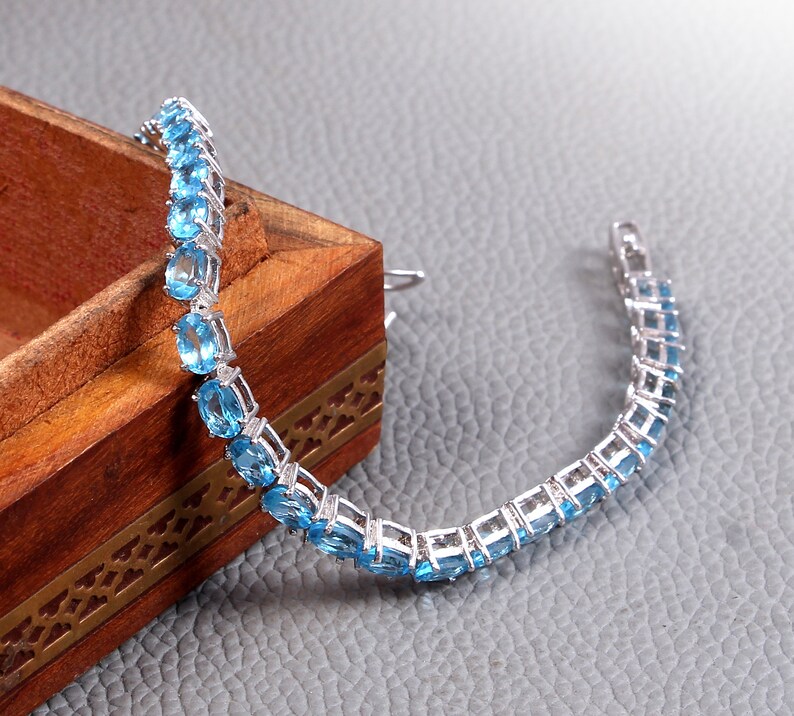 May include: A silver bracelet with a row of oval-shaped blue gemstones. The bracelet is lying on a wooden box with intricate carvings.