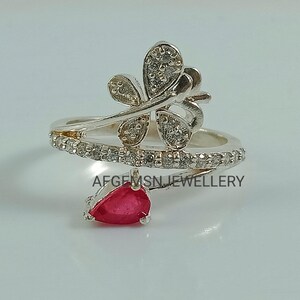 K&ouml;nnte beinhalten: Silberring mit einem Schmetterlingsdesign und einem roten Edelstein. Der Schmetterling ist mit klaren Steinen verziert und der Ring ist mit einer Reihe kleinerer klarer Steine verziert.