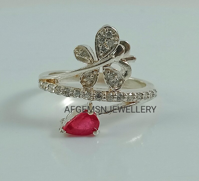 K&ouml;nnte beinhalten: Silberring mit einem Schmetterlingsdesign und einem roten Edelstein. Der Schmetterling ist mit klaren Steinen verziert und der Ring ist mit einer Reihe kleinerer klarer Steine verziert.