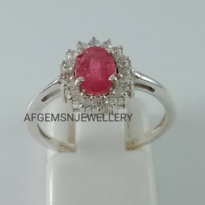 Pu&ograve; includere: Un anello d'argento con una pietra ovale rossa circondata da pietre bianche pi&ugrave; piccole. L'anello &egrave; incastonato in stile halo e ha una fascia semplice. Il testo "AFGEMSNJEWELLERY" &egrave; visibile sulla fascia.