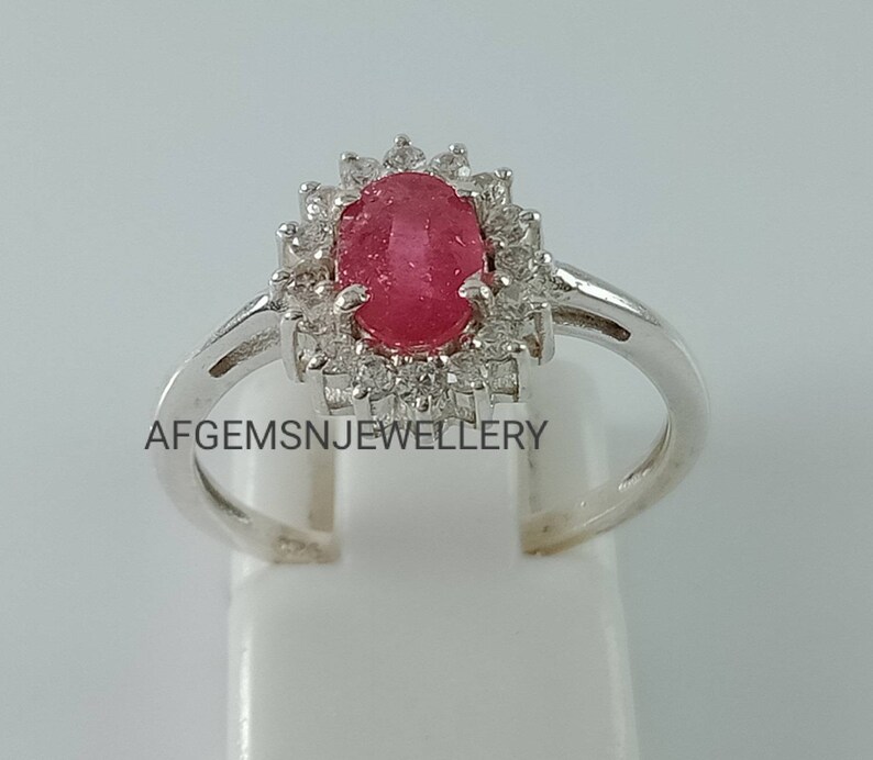 Pu&ograve; includere: Un anello d'argento con una pietra ovale rossa circondata da pietre bianche pi&ugrave; piccole. L'anello &egrave; incastonato in stile halo e ha una fascia semplice. Il testo "AFGEMSNJEWELLERY" &egrave; visibile sulla fascia.