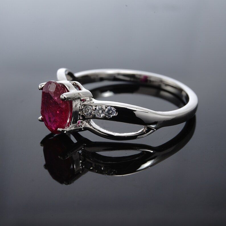 Pode incluir: Um anel de prata com uma grande pedra de rubi oval e pedras menores transparentes na banda.