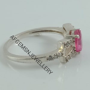 May include: A silver ring featuring a pink oval-shaped gemstone surrounded by smaller clear gemstones. The ring is displayed against a white backdrop.
