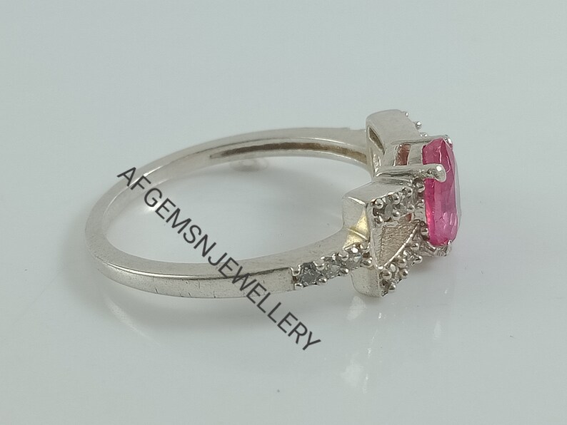 May include: A silver ring featuring a pink oval-shaped gemstone surrounded by smaller clear gemstones. The ring is displayed against a white backdrop.