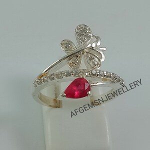 K&ouml;nnte beinhalten: Silberring mit einem roten Edelstein und einem Schmetterlingsdesign. Der Schmetterling hat klare Steine auf seinen Fl&uuml;geln. Der Ring liegt auf einem wei&szlig;en Hintergrund. AFGEMSNJEWELLERY ist auf dem Hintergrund geschrieben.
