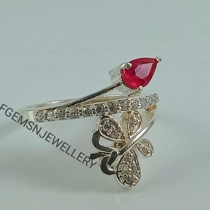 K&ouml;nnte beinhalten: Ein silberner Ring mit einem roten birnenf&ouml;rmigen Edelstein und einem Schmetterlingsdesign mit klaren Steinen.