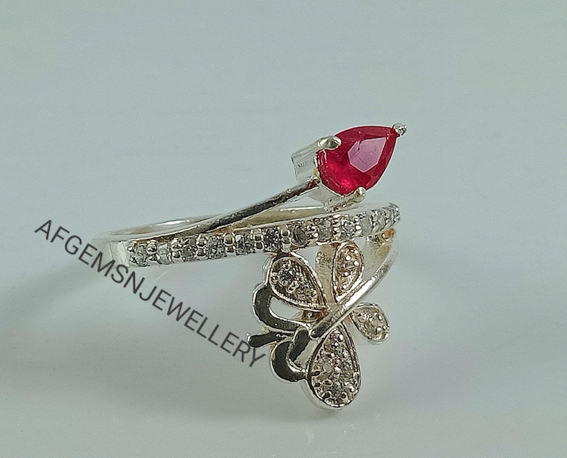 K&ouml;nnte beinhalten: Ein silberner Ring mit einem roten birnenf&ouml;rmigen Edelstein und einem Schmetterlingsdesign mit klaren Steinen.