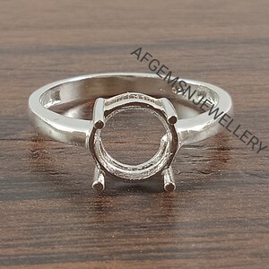 May include: A silver ring with a round setting for a gemstone. The ring is on a wooden surface.