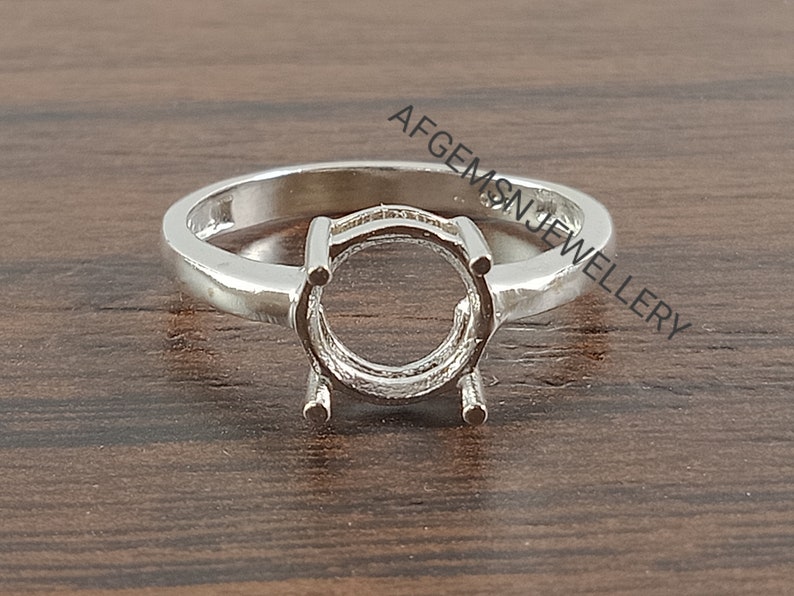 May include: A silver ring with a round setting for a gemstone. The ring is on a wooden surface.