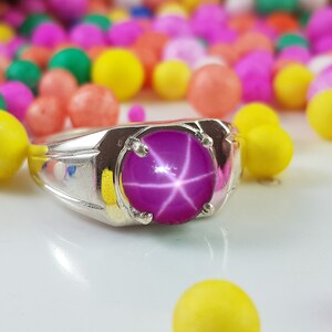 Op de afbeelding: Een zilveren ring met een grote, ronde, roze ster-saffier edelsteen. De edelsteen heeft een stervormig patroon in het midden.