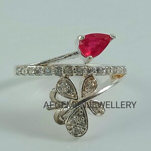 K&ouml;nnte beinhalten: Ein silberner Ring mit einem roten Edelstein und einem Schmetterlingsdesign. Der Schmetterling ist mit klaren Steinen besetzt, und der Ring ist mit klaren Steinen besetzt.