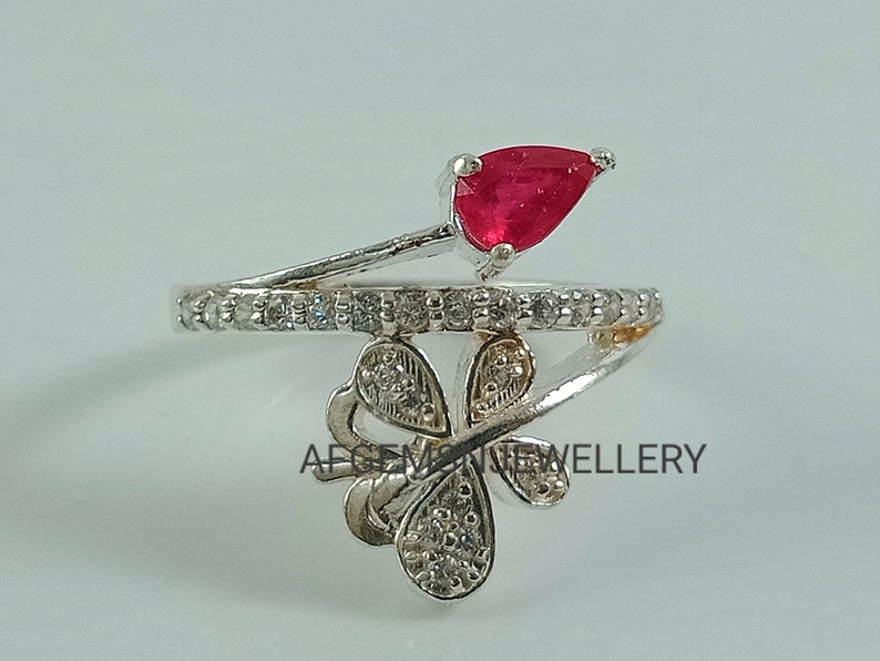 K&ouml;nnte beinhalten: Ein silberner Ring mit einem roten Edelstein und einem Schmetterlingsdesign. Der Schmetterling ist mit klaren Steinen besetzt, und der Ring ist mit klaren Steinen besetzt.