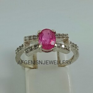 May include: A silver ring featuring a pink oval gemstone surrounded by smaller clear gemstones. The ring has a delicate design with a criss-cross pattern.