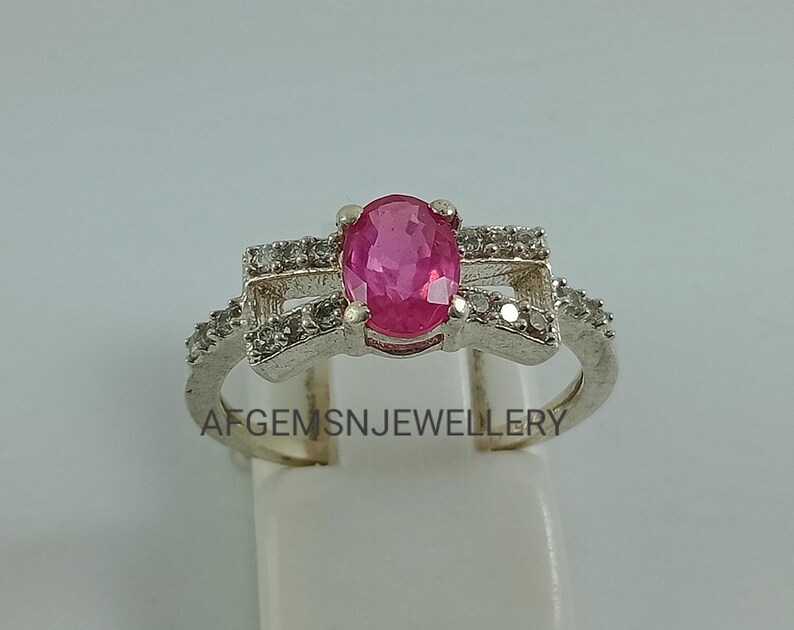 May include: A silver ring featuring a pink oval gemstone surrounded by smaller clear gemstones. The ring has a delicate design with a criss-cross pattern.