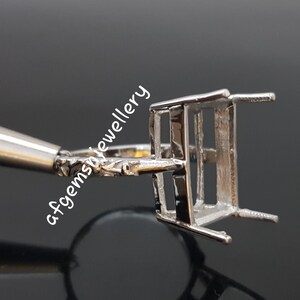 May include: Silver ring setting with a rectangular bezel for a gemstone. The setting has a delicate, ornate band.