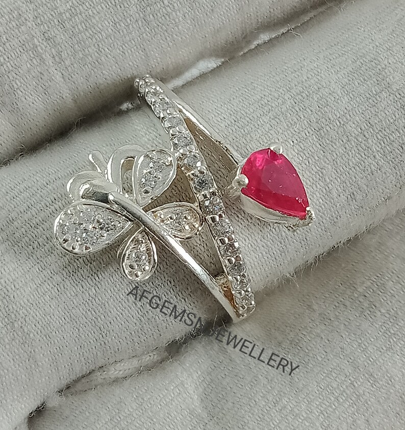 K&ouml;nnte beinhalten: Ein silberner Ring mit einem Schmetterlingsdesign und einem roten herzf&ouml;rmigen Stein. Der Ring ist mit klaren Zirkonia-Steinen besetzt.