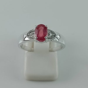 Könnte beinhalten: Ein silberner Ring mit einem roten ovalen Edelstein, der von kleineren weißen Edelsteinen umgeben ist.