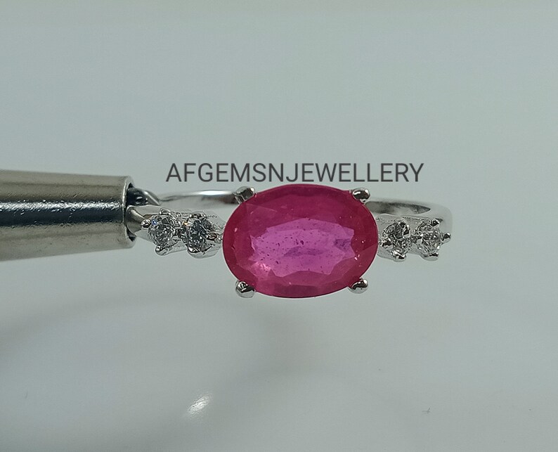 K&ouml;nnte beinhalten: Ein silberner Ring mit einem gro&szlig;en ovalen rosafarbenen Edelstein und zwei kleineren runden wei&szlig;en Edelsteinen auf jeder Seite. Der Ring liegt auf einem wei&szlig;en Hintergrund. Der Text 'AFGEMSNJEWELLERY' ist oben in der Mitte des Bildes zu sehen.