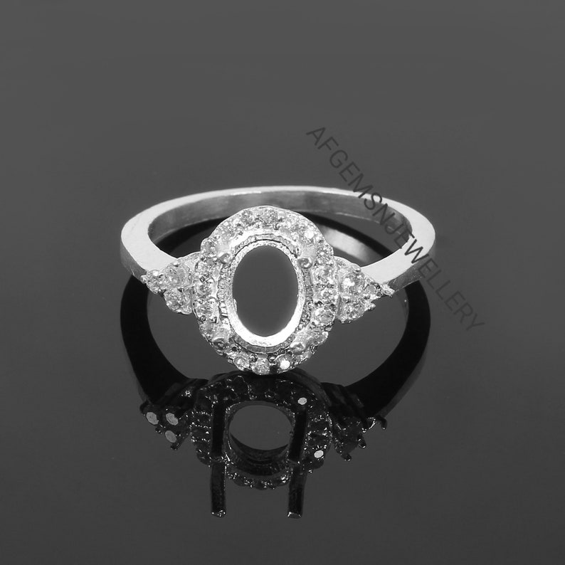 May include: Silver ring with an oval-shaped setting surrounded by small clear stones. The ring is set on a black background.