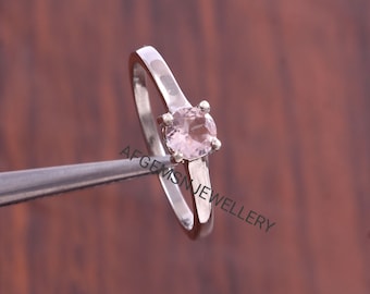 May include: A silver ring featuring a round, pale pink gemstone. The ring is held by tweezers. The text "AFGEMSNJEWELLERY" is visible on the ring.