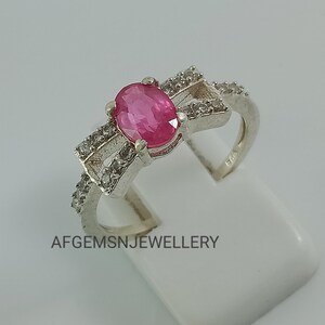 May include: A silver ring featuring a pink oval gemstone surrounded by smaller white gemstones. The ring has a delicate bow design and is hallmarked "925".