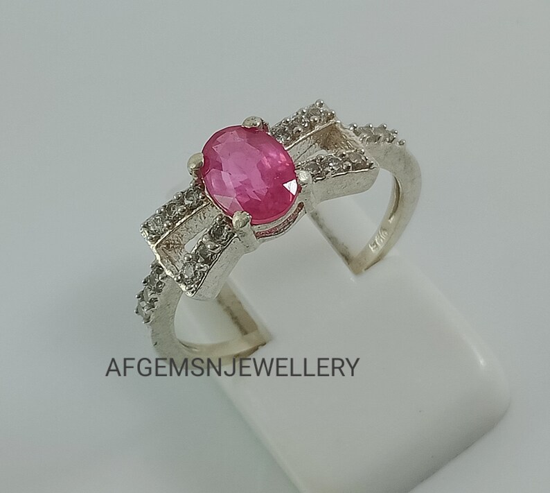 May include: A silver ring featuring a pink oval gemstone surrounded by smaller white gemstones. The ring has a delicate bow design and is hallmarked "925".