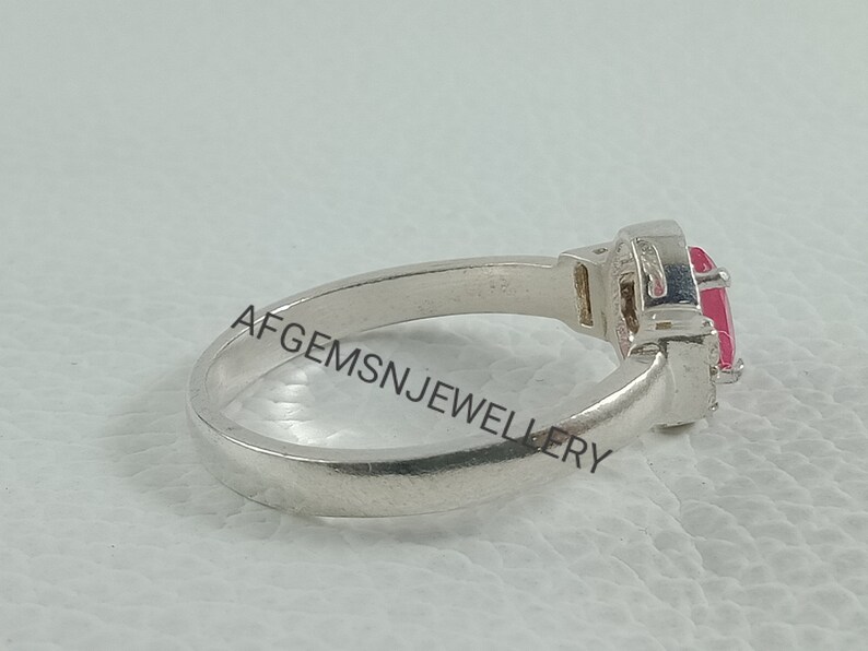 K&ouml;nnte beinhalten: Ein silberner Ring mit einem rosafarbenen Edelstein, der in einer Krappenfassung sitzt. Der Ring liegt auf einer wei&szlig;en Oberfl&auml;che.