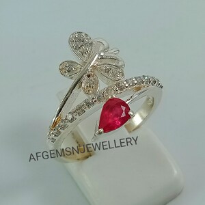 K&ouml;nnte beinhalten: Ein silberner Ring mit einem roten Edelstein und einem Schmetterlingsdesign. Der Schmetterling ist mit kleinen wei&szlig;en Steinen verziert.