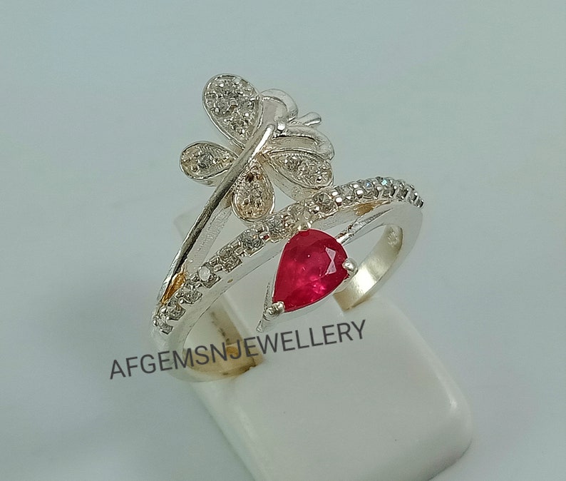 K&ouml;nnte beinhalten: Ein silberner Ring mit einem roten Edelstein und einem Schmetterlingsdesign. Der Schmetterling ist mit kleinen wei&szlig;en Steinen verziert.