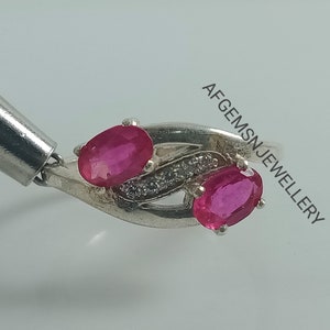 May include: A silver ring with two oval-shaped ruby gemstones and a row of smaller clear gemstones. The ring is set in a delicate, twisted design.