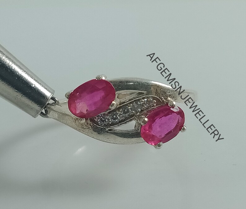 May include: A silver ring with two oval-shaped ruby gemstones and a row of smaller clear gemstones. The ring is set in a delicate, twisted design.