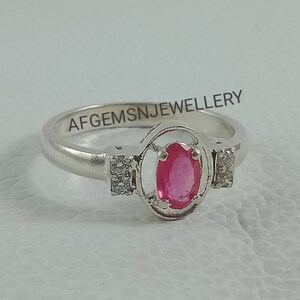 K&ouml;nnte beinhalten: Ein silberner Ring mit einem rosafarbenen ovalen Edelstein, der von einem silbernen Rahmen umgeben ist. Der Ring hat zwei kleine klare Edelsteine ​​auf beiden Seiten des Rahmens. Der Ring ist mit dem Text "AFGEMSNJEWELLERY" graviert.