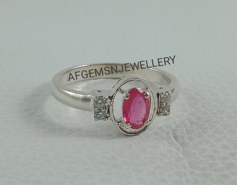 K&ouml;nnte beinhalten: Ein silberner Ring mit einem rosafarbenen ovalen Edelstein, der von einem silbernen Rahmen umgeben ist. Der Ring hat zwei kleine klare Edelsteine ​​auf beiden Seiten des Rahmens. Der Ring ist mit dem Text "AFGEMSNJEWELLERY" graviert.