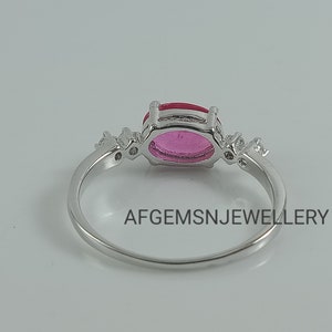 K&ouml;nnte beinhalten: Ein silberner Ring mit einem rosafarbenen ovalen Edelstein, der von kleineren klaren Edelsteinen umgeben ist. Der Ring ist in einer Krappenfassung gefasst und hat eine polierte Oberfl&auml;che. Der Text "AFGEMSNJEWELLERY" ist auf der Ringschiene sichtbar.