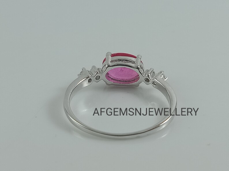 K&ouml;nnte beinhalten: Ein silberner Ring mit einem rosafarbenen ovalen Edelstein, der von kleineren klaren Edelsteinen umgeben ist. Der Ring ist in einer Krappenfassung gefasst und hat eine polierte Oberfl&auml;che. Der Text "AFGEMSNJEWELLERY" ist auf der Ringschiene sichtbar.