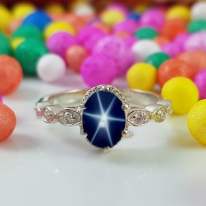 May include: A silver ring with a large, oval, dark blue gemstone displaying a star-like effect. The ring is accented with small, clear stones and decorative detailing. Colorful spheres of various sizes and hues form the background.