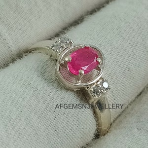 K&ouml;nnte beinhalten: Ein silberner Ring mit einem rosafarbenen ovalen Edelstein, der von kleineren wei&szlig;en Edelsteinen umgeben ist. Der Ring ist in einem filigranen, durchbrochenen Design gefasst.