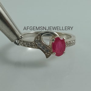 May include: A silver ring with a heart design and a red oval gemstone. The ring is set with small white gemstones around the band.