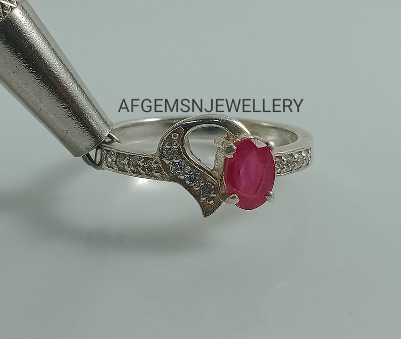 May include: A silver ring with a heart design and a red oval gemstone. The ring is set with small white gemstones around the band.