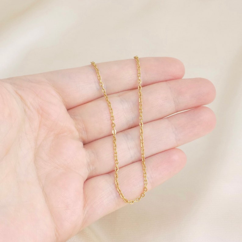 May include: A gold chain necklace with a delicate, thin chain link design. The chain is shown lying on a person's hand.