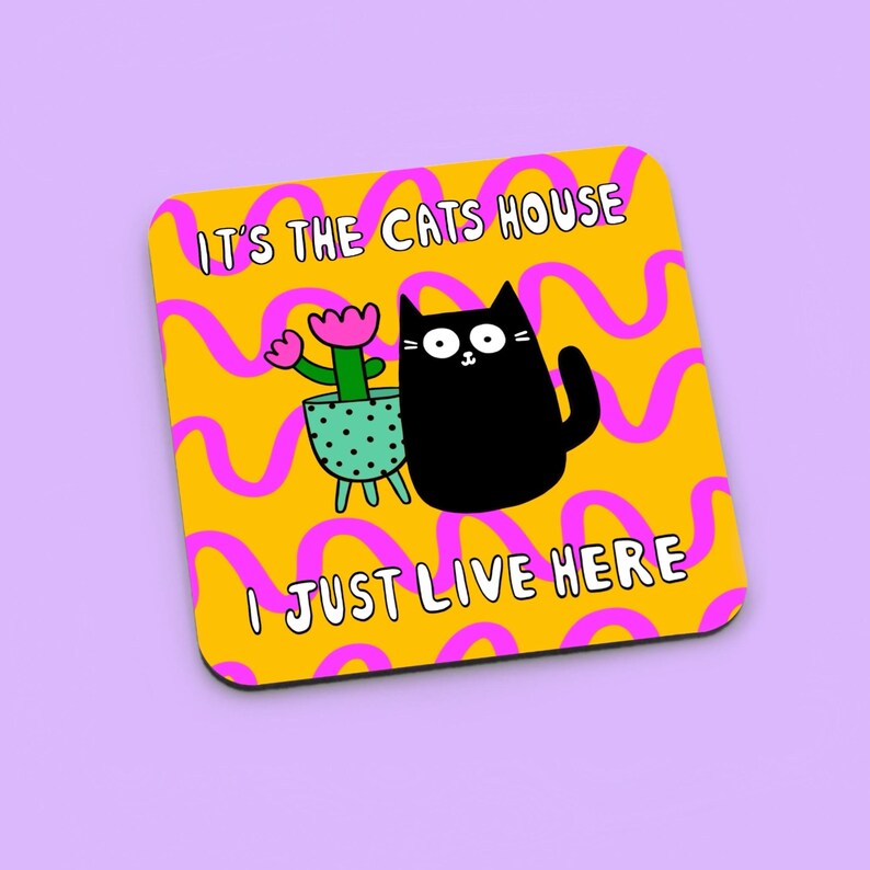 May include: A yellow coaster with pink wavy lines and the text "It's the cats house I just live here". A black cat with white eyes and a pink flower in a green pot are in the center of the coaster.