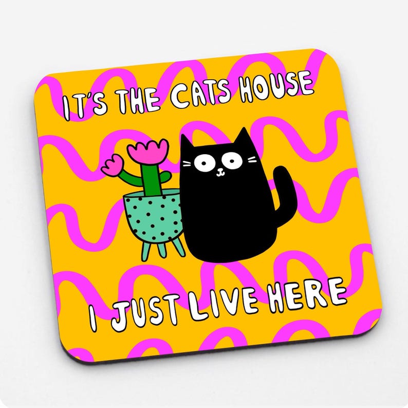 May include: A yellow and pink coaster with a black cat and a green potted plant. The text on the coaster reads "It's the cats house I just live here."