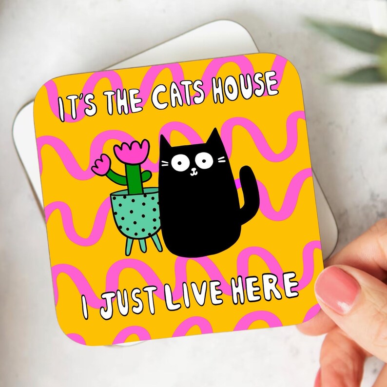 May include: A yellow and pink coaster with a black cat and a potted plant. The text on the coaster reads "It's the cats house I just live here."