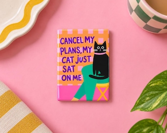 May include: A rectangular magnet featuring a black cat sitting on a person's lap. The background is an orange and pink checkered pattern. The text in purple reads: CANCEL MY PLANS, MY CAT JUST SAT ON ME. The design includes teal legs and a yellow stool.