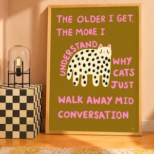 May include: A framed rectangular wall art print with a dark olive green background. It features a central illustration of a cream-colored cat with black spots and the pink text: THE OLDER I GET, THE MORE I UNDERSTAND WHY CATS JUST WALK AWAY MID CONVERSATION.