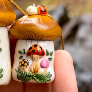 May include: A set of three small ceramic mushroom ornaments with brown and white caps and white stems. The mushrooms are decorated with red and black spots, yellow and black spots, and pink dots. The ornaments are strung together with twine.