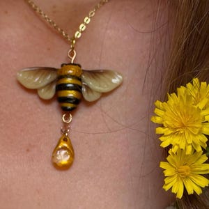 May include: A gold-colored necklace with a bee pendant. The bee pendant has a yellow and black striped body, translucent wings, and a teardrop-shaped charm. Yellow flowers are in the background.