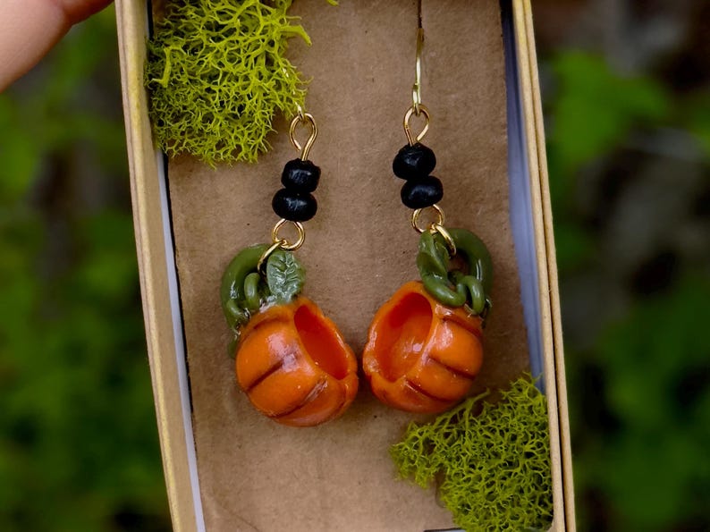 May include: A pair of dangle earrings featuring miniature pumpkin charms with green stems and black bead accents. The pumpkins are orange and have a textured surface.