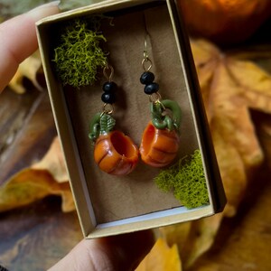 May include: A pair of pumpkin earrings with gold hooks and black beads. The pumpkins are orange and green and are in a small box with moss.