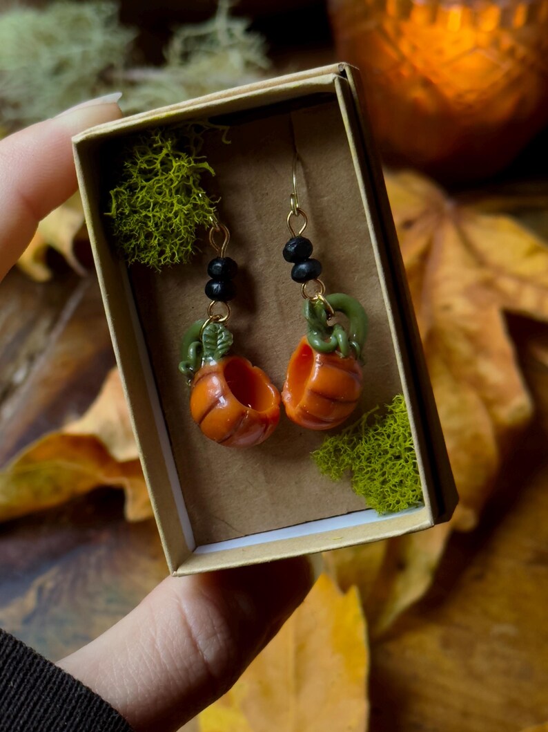 May include: A pair of pumpkin earrings with gold hooks and black beads. The pumpkins are orange and green and are in a small box with moss.